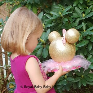 May include: A gold and pink Minnie Mouse themed centerpiece with a tulle skirt and a crown base. The centerpiece is made of two large gold glitter balls with pink polka dot ears and a pink bow.