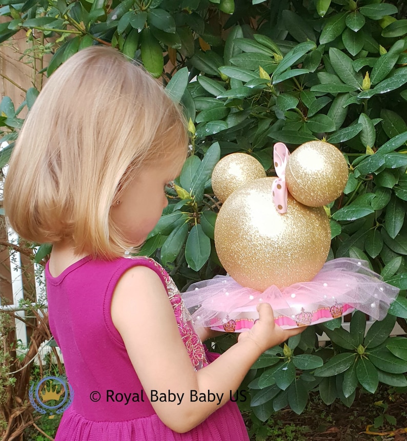 May include: A gold and pink Minnie Mouse themed centerpiece with a tulle skirt and a crown base. The centerpiece is made of two large gold glitter balls with pink polka dot ears and a pink bow.