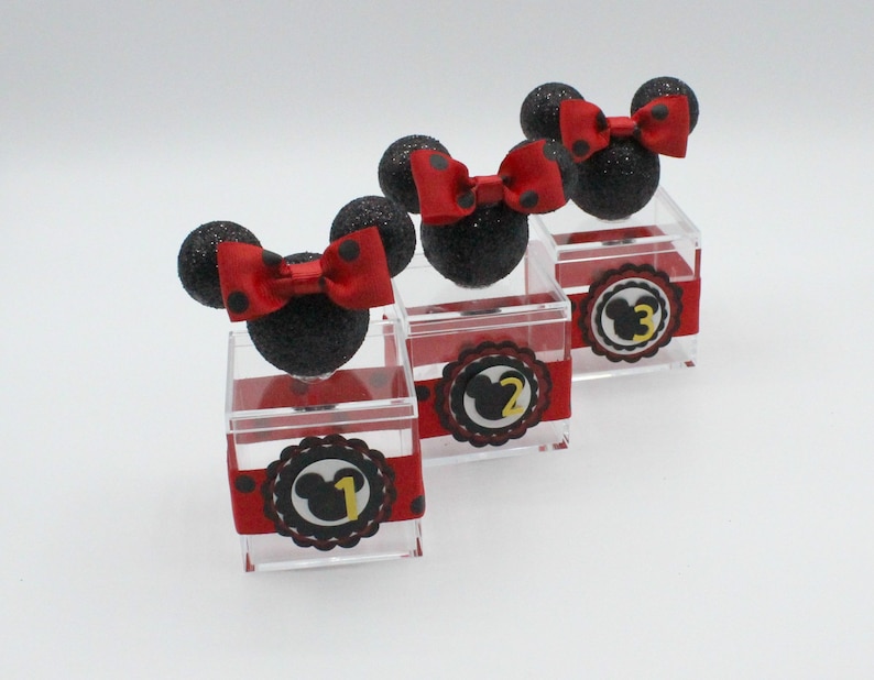 K&ouml;nnte beinhalten: Drei transparente Acrylboxen mit schwarzen und roten Minnie Mouse Ohren und Schleifen. Jede Box hat ein schwarz-rotes gepunktetes Band und einen schwarzen und gelben Kreis mit einem Minnie Mouse Kopf und der Nummer 1, 2 oder 3.
