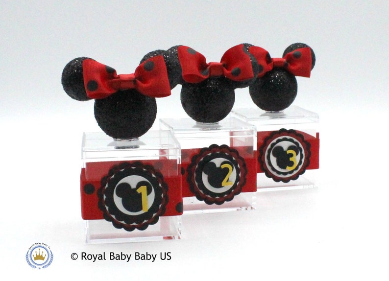 K&ouml;nnte beinhalten: Drei transparente Acrylboxen mit schwarzen und roten Minnie Mouse Ohren und Schleifen oben. Jede Box hat einen schwarzen und roten Kreis mit einem wei&szlig;en Mickey Mouse Kopf und einer goldenen Zahl darin. Die Zahlen sind 1, 2 und 3.