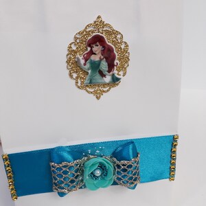 Może przedstawiać: Biała torba na prezent ozdobiona turkusową wstążką i kokardą, ozdobiona złotymi wykończeniami i turkusowym kwiatem. Złota brokatowa ramka ze zdjęciem Arielki z Małej Syrenki jest przymocowana do torby.