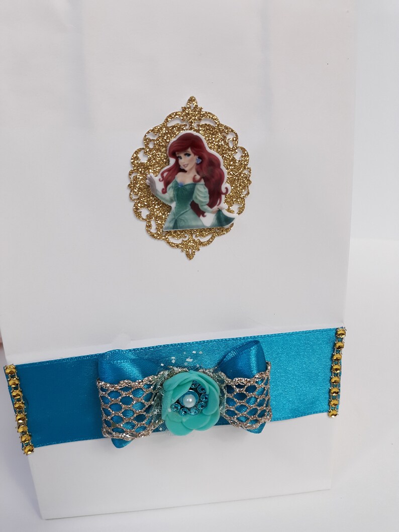 Może przedstawiać: Biała torba na prezent ozdobiona turkusową wstążką i kokardą, ozdobiona złotymi wykończeniami i turkusowym kwiatem. Złota brokatowa ramka ze zdjęciem Arielki z Małej Syrenki jest przymocowana do torby.