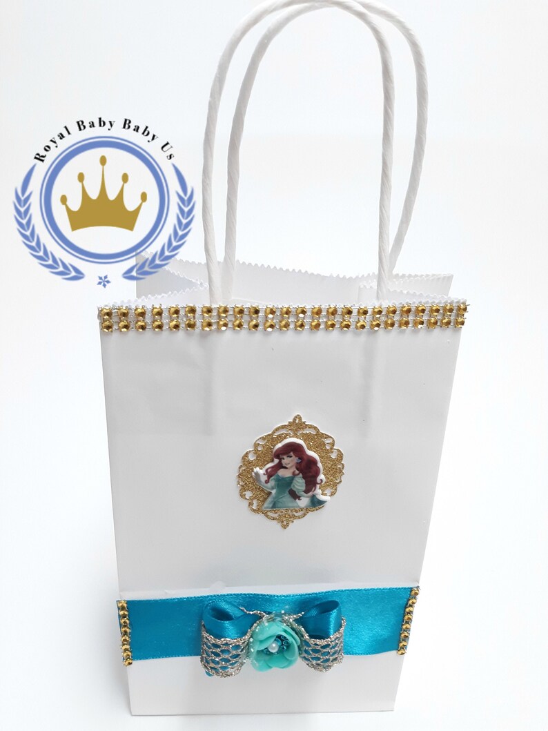 Może przedstawiać: Biała torba na prezent ze złotym obszyciem z dżet&oacute;w i turkusową kokardą. Zawiera złote zdjęcie Arielki z Małej Syrenki. Torba ma białe uchwyty ze sznurka i tekst "Royal Baby Baby Us" w okrągłym logo.