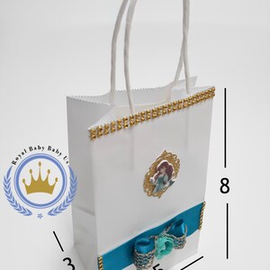 Może przedstawiać: Biała torba na prezent z turkusową wstążką i kokardą oraz złotym wykończeniem. Torba przedstawia obraz Arielki z Małej Syrenki. Torba ma 20 cm wysokości, 13 cm szerokości i 8 cm głębokości.