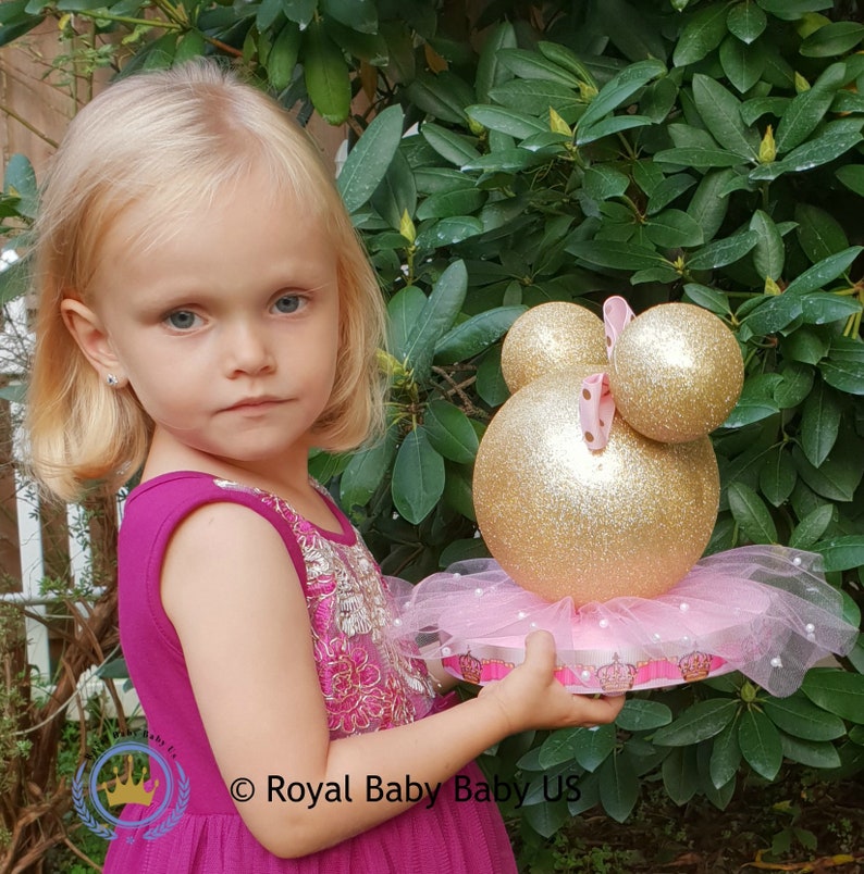 May include: A gold and pink Minnie Mouse themed centerpiece with a tulle skirt and a crown on the base.  The centerpiece is held by a young child.