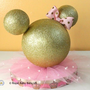 May include: Gold glitter Mickey Mouse ears centerpiece with a pink tulle skirt and a pink bow with white polka dots.  The base is decorated with a pink and gold ribbon with a crown pattern.