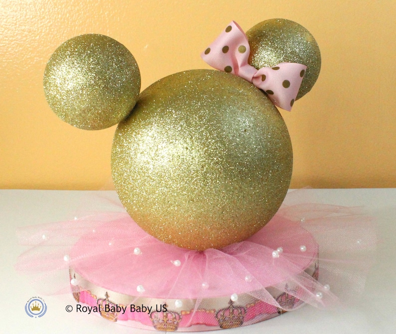 May include: Gold glitter Mickey Mouse ears centerpiece with a pink tulle skirt and a pink bow with white polka dots.  The base is decorated with a pink and gold ribbon with a crown pattern.