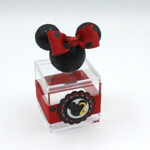 K&ouml;nnte beinhalten: Eine transparente Plastikbox mit einem schwarzen und roten Minnie Mouse Kopf oben drauf. Die Box hat ein rotes Band mit wei&szlig;en Punkten und einen schwarzen und wei&szlig;en Kreis mit einer gelben Zahl 2 in der Mitte.