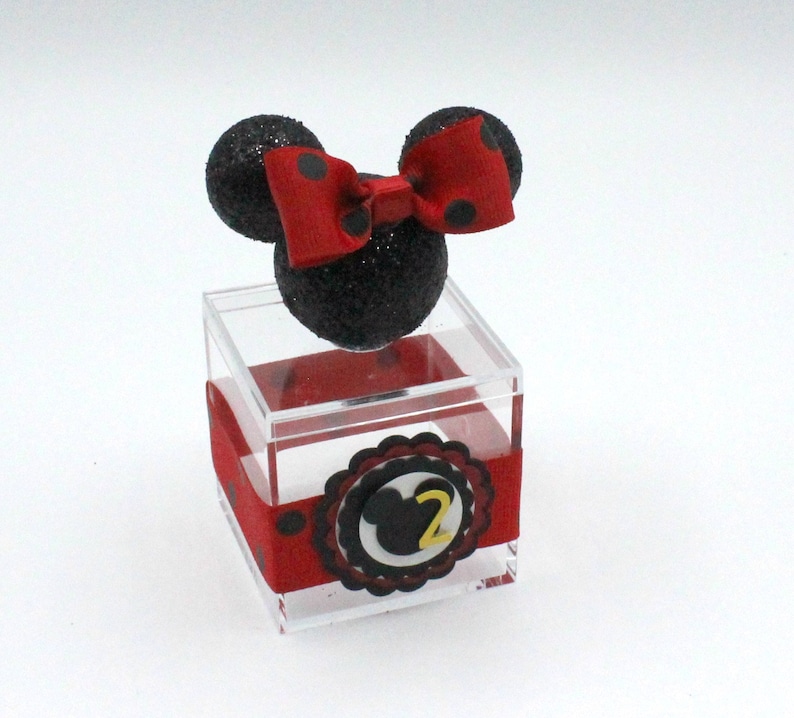 K&ouml;nnte beinhalten: Eine transparente Plastikbox mit einem schwarzen und roten Minnie Mouse Kopf oben drauf. Die Box hat ein rotes Band mit wei&szlig;en Punkten und einen schwarzen und wei&szlig;en Kreis mit einer gelben Zahl 2 in der Mitte.
