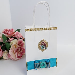 Może przedstawiać: Biała torba na prezent ze złotym wykończeniem i turkusową kokardą. Torba posiada oprawiony obrazek postaci z kresk&oacute;wki. Kokarda ma ozdobny guzik pośrodku. Torba ma białe skręcone uchwyty.