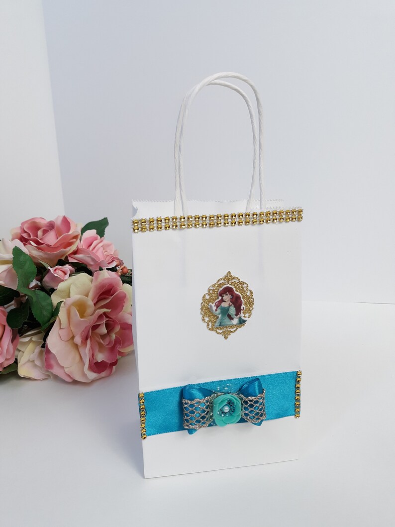 Może przedstawiać: Biała torba na prezent ze złotym wykończeniem i turkusową kokardą. Torba posiada oprawiony obrazek postaci z kresk&oacute;wki. Kokarda ma ozdobny guzik pośrodku. Torba ma białe skręcone uchwyty.