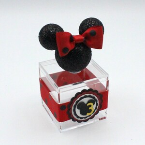 K&ouml;nnte beinhalten: Eine transparente Acrylbox mit einem rot-schwarz gepunkteten Band und einem schwarz-roten Minnie Mouse Kopf mit einer roten Schleife oben. Die Box hat einen wei&szlig;en Kreis mit einem schwarzen Minnie Mouse Kopf und der Zahl 3 in Gelb.