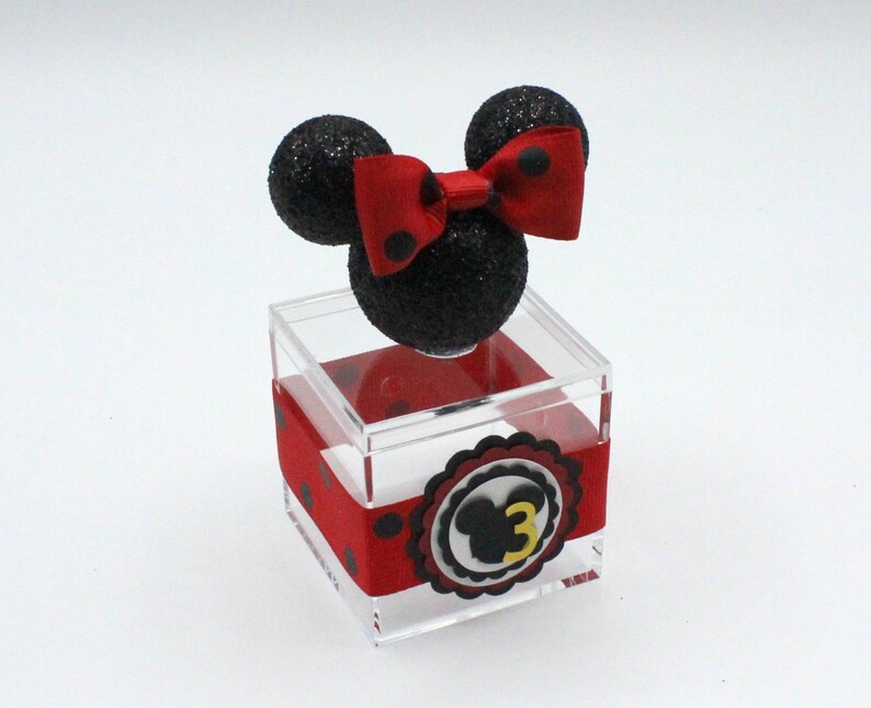 K&ouml;nnte beinhalten: Eine transparente Acrylbox mit einem rot-schwarz gepunkteten Band und einem schwarz-roten Minnie Mouse Kopf mit einer roten Schleife oben. Die Box hat einen wei&szlig;en Kreis mit einem schwarzen Minnie Mouse Kopf und der Zahl 3 in Gelb.