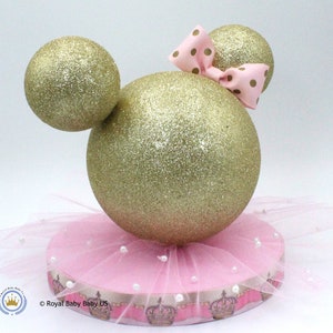 May include: Gold glitter Mickey Mouse ears centerpiece with a pink tulle skirt and a pink bow with gold polka dots. The base is pink with a white ribbon and gold crown accents. &copy; Royal Baby US.