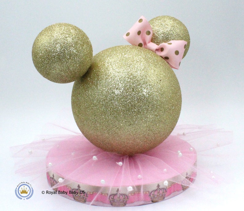 May include: Gold glitter Mickey Mouse ears centerpiece with a pink tulle skirt and a pink bow with gold polka dots. The base is pink with a white ribbon and gold crown accents. &copy; Royal Baby US.