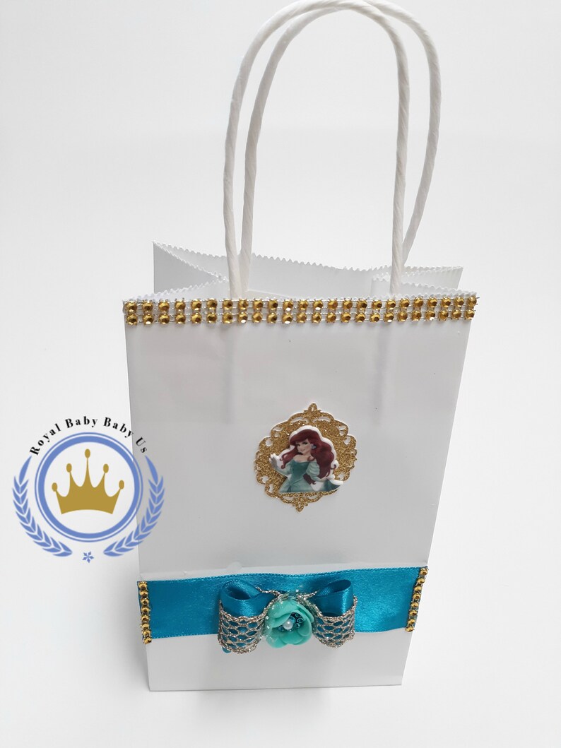 Może przedstawiać: Biała torba na prezent ze złotym wykończeniem i turkusową wstążką. Zawiera grafikę księżniczki i tekst "Royal Baby Baby Us". Zawiera białe papierowe uchwyty.