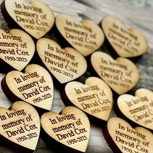 May include: Wooden heart-shaped memorial tokens with dark brown edges and light brown faces. Each heart is engraved with the text "In loving memory of David Cox 1950-2025". The hearts are arranged in a scattered pattern.