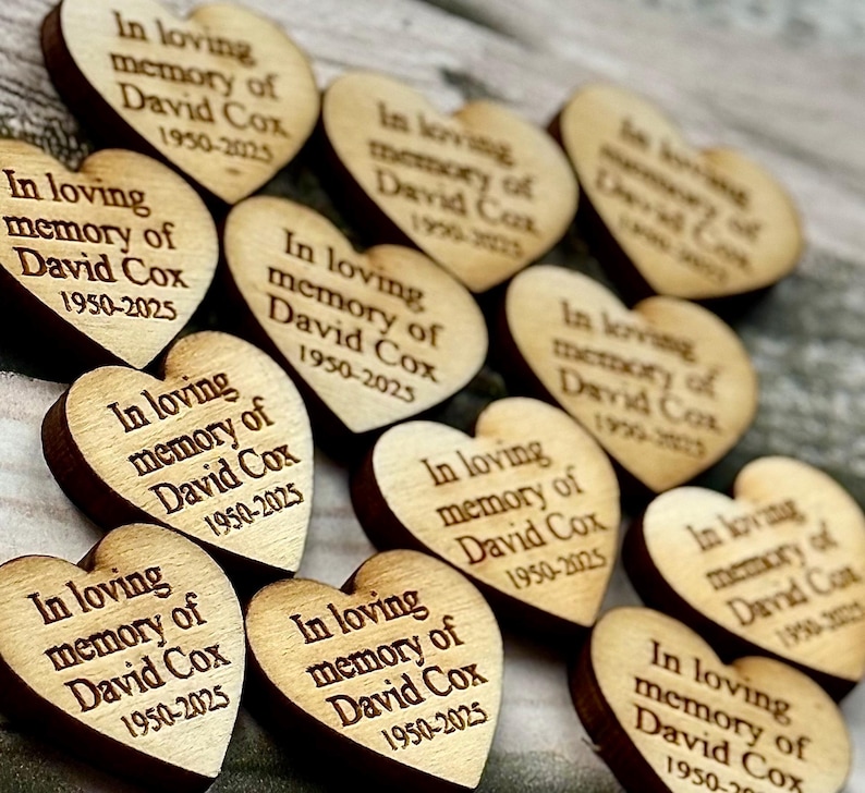 May include: Wooden heart-shaped memorial tokens with dark brown edges and light brown faces. Each heart is engraved with the text "In loving memory of David Cox 1950-2025". The hearts are arranged in a scattered pattern.