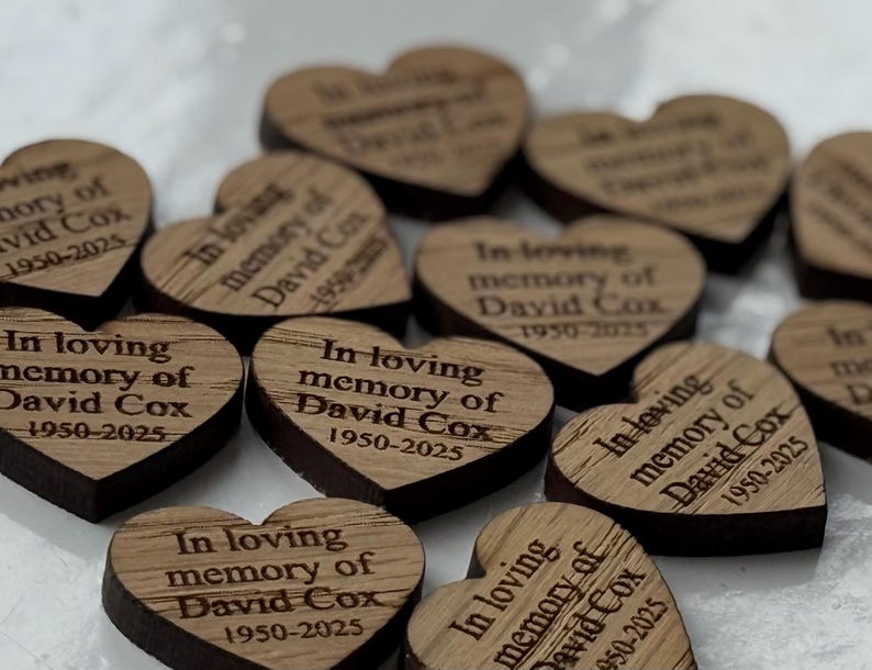May include: A collection of heart-shaped wooden tokens. Each token is engraved with the text "In loving memory of David Cox 1950-2025". The wood has a natural grain and a dark brown edge.