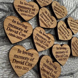 May include: A collection of heart-shaped wooden keepsakes. Each heart is engraved with the text "In loving memory of David Cox 1950-2025". The wooden hearts are arranged on a textured, silver surface.