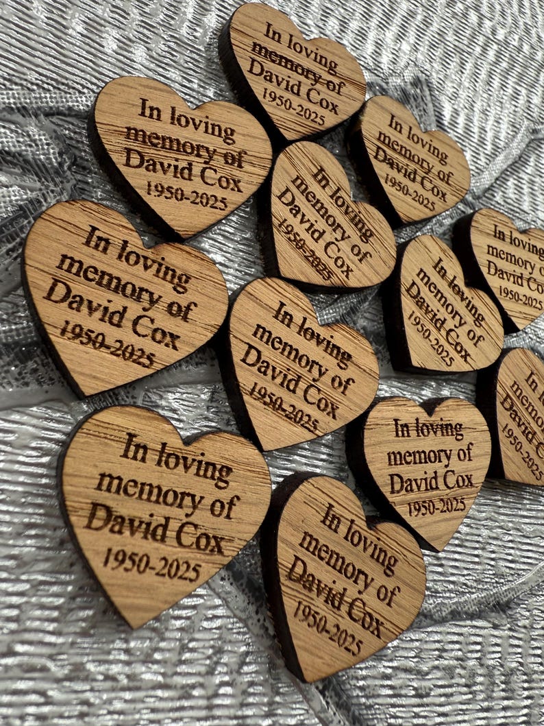 May include: A collection of heart-shaped wooden keepsakes. Each heart is engraved with the text "In loving memory of David Cox 1950-2025". The wooden hearts are arranged on a textured, silver surface.