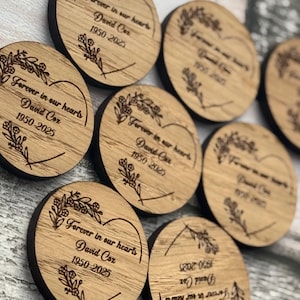 以下が含まれることがあります： 丸い木製のコースターのコレクション。各コースターは、テキスト「Forever in our hearts」、名前「David Cox」、および年「1950-2025」と花のアクセントが刻まれています。暗い茶色の縁があります。