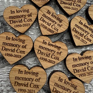 May include: A collection of heart-shaped wooden keepsakes. Each heart is engraved with the text "In loving memory of David Cox 1950-2025". The wood has a natural grain and a warm, light brown color.