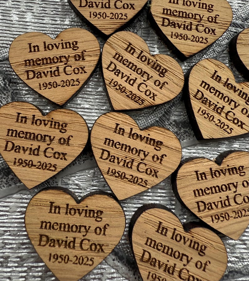 May include: A collection of heart-shaped wooden keepsakes. Each heart is engraved with the text "In loving memory of David Cox 1950-2025". The wood has a natural grain and a warm, light brown color.