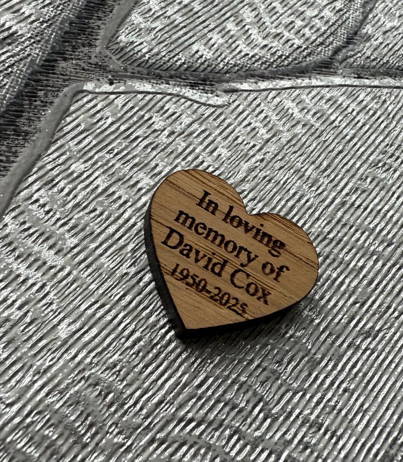 May include: A wooden heart-shaped memorial piece with the inscription "In loving memory of David Cox 1950-2025" is displayed. The text is engraved in a simple, readable font. The heart is made of light-colored wood.