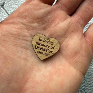 May include: A wooden heart-shaped memorial piece with the inscription "In loving memory of David Cox 1950-2025". The heart is light brown with visible wood grain and is held in a hand.