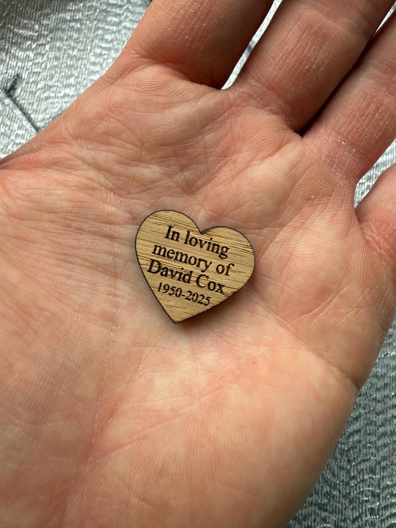 May include: A wooden heart-shaped memorial piece with the inscription "In loving memory of David Cox 1950-2025". The heart is light brown with visible wood grain and is held in a hand.
