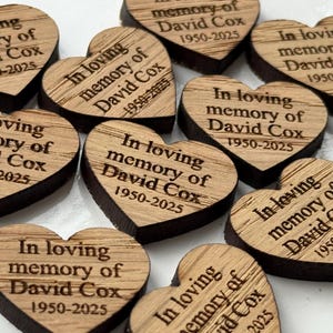 May include: Wooden heart-shaped memorial keepsakes. Each heart is engraved with the text "In loving memory of David Cox 1950-2025". The hearts have a light brown surface with dark brown edges.