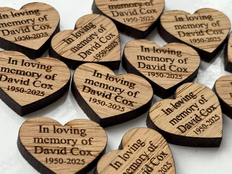 May include: Wooden heart-shaped memorial keepsakes. Each heart is engraved with the text "In loving memory of David Cox 1950-2025". The hearts have a light brown surface with dark brown edges.