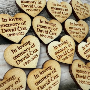 May include: Wooden heart-shaped memorial keepsakes. Each heart is engraved with the text "In loving memory of David Cox 1950-2025". The hearts are made of light-colored wood with a natural grain.