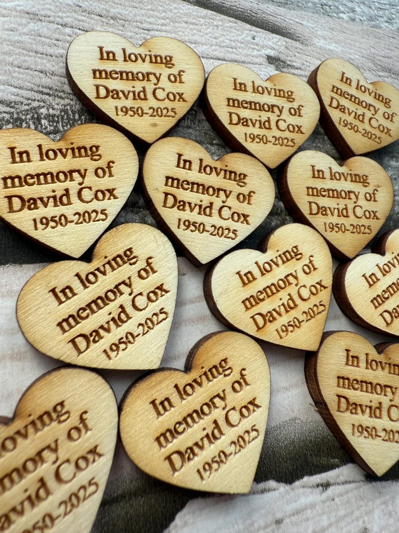 May include: Wooden heart-shaped memorial keepsakes. Each heart is engraved with the text "In loving memory of David Cox 1950-2025". The hearts are made of light-colored wood with a natural grain.