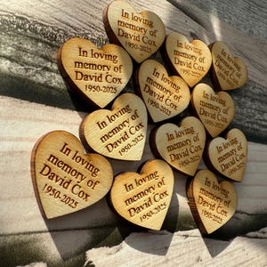 May include: A collection of wooden heart-shaped keepsakes. Each heart is engraved with the text "In loving memory of David Cox 1950-2025". The hearts are arranged on a light-colored wooden surface, creating a touching memorial.