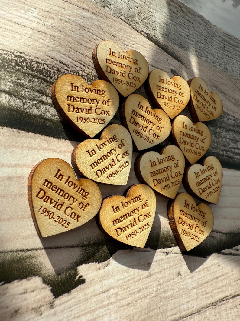 May include: A collection of wooden heart-shaped keepsakes. Each heart is engraved with the text "In loving memory of David Cox 1950-2025". The hearts are arranged on a light-colored wooden surface, creating a touching memorial.