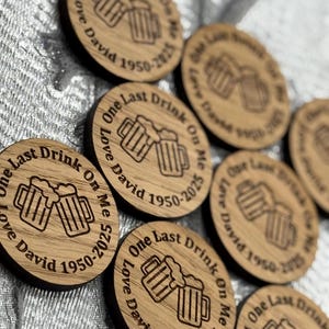 May include: A collection of round wooden coasters with laser-etched designs. Each coaster features two beer mugs and the text "One Last Drink On Me Love David 1950-2025". The coasters are made of light-colored wood.