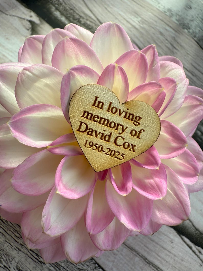 May include: A close-up of a pink and white dahlia flower with a wooden heart-shaped plaque. The plaque is engraved with the words "In loving memory of David Cox 1950-2025". The flower petals are soft and delicate.