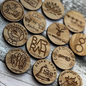 May include: A selection of circular wooden tokens, each engraved with unique designs and text. The tokens display phrases such as "The Rolling Barrel," "Cakes by Andrea," and "Drink Token." Decorative elements include a deer and a bell, all in a natural wood finish.