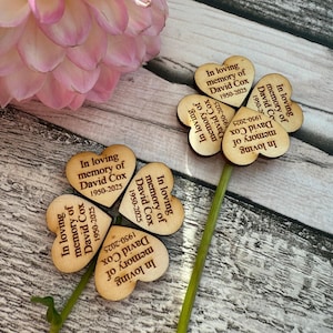 May include: Wooden heart-shaped memorial decorations with the text "In loving memory of David Cox 1950-2025". The decorations are arranged to resemble flowers, with green stems. A pink flower is in the background.