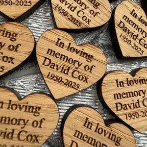 May include: Close-up of several wooden heart-shaped memorial keepsakes. Each heart is engraved with the text "In loving memory of David Cox 1950-2025". The wood has a natural grain and a dark border. A touching tribute.