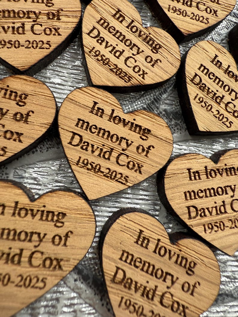 May include: Close-up of several wooden heart-shaped memorial keepsakes. Each heart is engraved with the text "In loving memory of David Cox 1950-2025". The wood has a natural grain and a dark border. A touching tribute.
