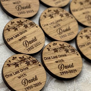 Könnte beinhalten: Eine Sammlung runder Holzuntersetzer mit dunkelbraunem Rand und hellbrauner Mitte. Jeder Untersetzer ist mit dem Text "One Last Drink with David 1950-2025" und einem floralen Design graviert.