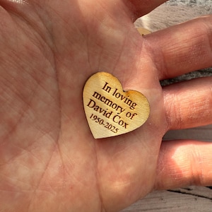May include: A small, heart-shaped wooden keepsake with the inscription "In loving memory of David Cox 1950-2025" engraved in a dark brown font. The heart is held in a hand, showcasing its natural wood grain.