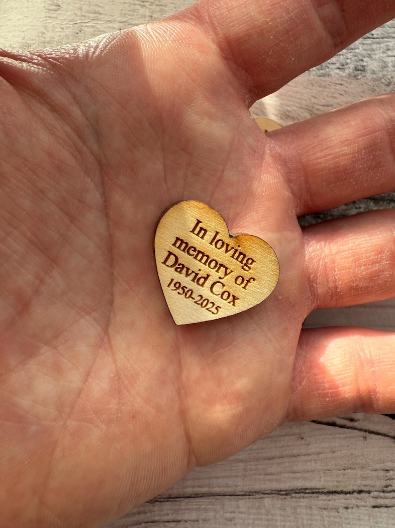 May include: A small, heart-shaped wooden keepsake with the inscription "In loving memory of David Cox 1950-2025" engraved in a dark brown font. The heart is held in a hand, showcasing its natural wood grain.