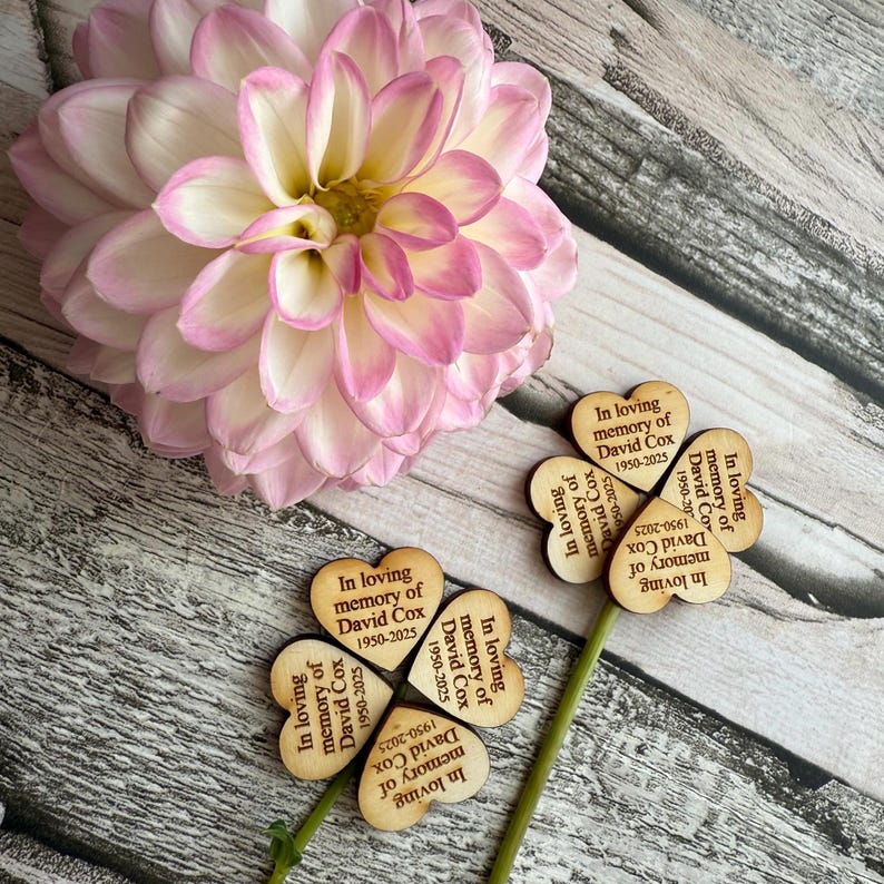 May include: A large pink and white dahlia flower with heart-shaped wooden memorial stakes. The stakes read "In loving memory of David Cox 1950-2025". The flower and stakes are on a weathered wood surface.