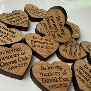 May include: A collection of heart-shaped wooden keepsakes. Each heart is engraved with the text "In loving memory of David Cox 1950-2025". The wood has a natural grain and a dark brown edge.