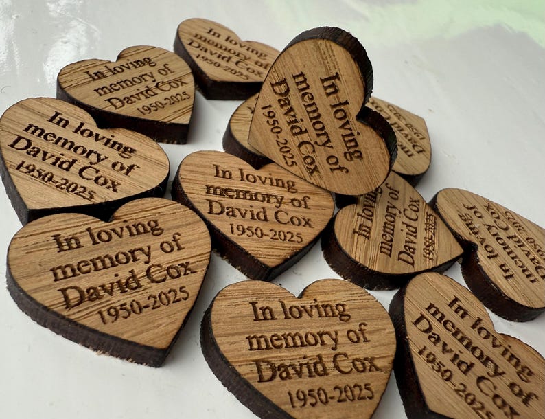 May include: A collection of heart-shaped wooden keepsakes. Each heart is engraved with the text "In loving memory of David Cox 1950-2025". The wood has a natural grain and a dark brown edge.
