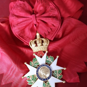 Puede incluir: Primer plano de una cinta roja y una corona dorada. La cinta está atada en un lazo. Debajo, una medalla en forma de estrella blanca y dorada con un retrato de un hombre y la inscripción "NAPOLEON EMP. DES FRANCAIS".
